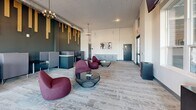 Located at 100 Foundry Dr in West Lafayette, Indiana, this thoughtfully designed single-floor workspace blends collaborative and focused work environments into one cohesive layout. The space is anchored by an open lounge area furnished with contemporary burgundy pod-style chairs and sleek glass-top coffee tables, creating an inviting atmosphere for casual meetings or relaxed work sessions. A striking dark-toned accent wall serves as a dramatic backdrop, adorned with cascading vertical wood slat installations that add warmth and an artistic focal point. Pendant chandeliers and exposed ductwork contribute an industrial-modern aesthetic to the ceiling. Along the perimeter, a row of semi-private work pods with built-in desks and partitioned panels provides quiet, individual workstations ideal for focused tasks. Large windows flood the interior with natural light and offer views of the surrounding outdoor landscape. Near the entrance, double glass doors open into a welcoming reception-style foyer featuring built-in cabinetry and elevated transom windows. Mounted displays and a statement wall clock add functional and decorative character throughout. Patterned carpet flooring ties the entire space together, completing a professional yet comfortable environment suited for productivity and collaboration.