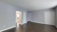 This single-floor space presents a clean, move-in-ready interior defined by a cohesive and understated aesthetic. The main living area immediately draws attention with its rich, dark hardwood flooring that contrasts elegantly against the soft blue-gray walls and crisp white baseboards and trim. The neutral color palette creates a calm, versatile atmosphere that adapts easily to a variety of furnishing styles. The room is open and well-proportioned, offering a generous sense of space with smooth, white ceilings that amplify the brightness of the interior. A classic six-panel white door provides access to an exterior entry point, while an open doorway connects seamlessly to an adjacent room, hinting at a thoughtful and functional layout. A closet with a white paneled door adds practical storage directly within the main living area. The adjoining room, visible through the open passage, continues the same warm, light-filled character with matching flooring and a baseboard heating unit along the wall. Throughout the space, the consistent use of white trim, panel doors, and cool-toned wall color ties each area together into a unified and polished whole, making it an inviting and adaptable environment.