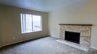This single-floor space centers around a comfortable living area that combines functional design with warm, inviting details. The room is finished with wall-to-wall neutral carpeting, which adds softness underfoot and complements the light beige walls and white ceiling. A standout feature is the stacked stone fireplace, clad in layered ledger-style stonework in warm tan and gray tones, topped with a natural wood mantel. The raised tile hearth provides a clean, defined transition from the carpeted floor. Adjacent to the fireplace, a large picture window fills the room with natural light and offers views of surrounding trees and neighboring rooftops, creating a pleasant indoor-outdoor connection. The window's vertical framing adds architectural interest while maximizing the light entering the space. The overall layout is open and uncluttered, allowing for flexible furniture arrangement. The neutral color palette throughout—from the flooring to the walls—creates a calm, cohesive ambiance that would suit a variety of interior styles. The combination of the stone fireplace and generous natural light makes this living space both practical and characterful.