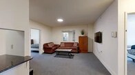 Located at 327 Eddy St in Ithaca, NY, this five-bedroom, two-bathroom unit is thoughtfully laid out across a single floor, offering a functional and comfortable shared living arrangement. The open living area is carpeted and furnished with a sofa set, wall-mounted TV, and a tile-topped coffee table, with a wooden storage cabinet adding warmth to the space. A pass-through window connects the living area to the kitchen, which features dark granite countertops, a breakfast bar with bar stools, white appliances including a refrigerator, range, and microwave, and a bold red backsplash that adds a pop of color. The two full bathrooms are finished with light tile flooring, gray cabinetry, undermount sinks with granite countertops, and a combination tub and shower with sliding glass doors. Each of the five bedrooms is carpeted and features neutral walls, with several rooms offering windows that bring in natural light and views of the surrounding area. Some bedrooms include individual wall-unit climate control. Connecting hallways provide easy access throughout the unit, making the layout well-suited for multiple occupants. This unit offers a practical and well-appointed living space in the heart of Ithaca.