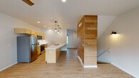 This three-story home showcases a thoughtful blend of modern design and functional living across multiple levels. The ground floor houses a spacious attached two-car garage, equipped with an automatic opener and baseboard heating, providing ample storage and parking. The main living level centers around an open-concept kitchen and living area. The kitchen is well-appointed with warm honey-toned cabinetry, stainless steel appliances including a refrigerator, range, and microwave, and a generous island featuring a striking quartz countertop with subtle veining and an integrated sink. Pendant lighting in an industrial style hangs above the island, while recessed lighting brightens the space throughout. A standout architectural feature is the floor-to-ceiling bamboo-paneled accent wall, which adds warmth and visual interest. Light wood-tone flooring flows seamlessly throughout the main level. An upper living area includes a bright, open room with sliding glass doors leading to a private balcony, offering outdoor views and natural light, complemented by a ceiling fan overhead. Bedrooms on the upper floors include walk-in closets with built-in shelving. Bathrooms feature tile flooring, wood-toned vanities with white countertops, and tub-shower combinations, offering a clean and consistent aesthetic throughout the home.