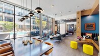 This expansive single-floor amenity lounge is a thoughtfully designed communal space that blends bold contemporary style with a wide range of social and recreational functions. Floor-to-ceiling glass walls line the perimeter, flooding the interior with abundant natural light and providing sweeping views of the resort-style outdoor pool courtyard, complete with lounge chairs, lush landscaping, and an outdoor grilling station.

The interior is divided into several distinct yet open zones. A bright lounge area features large sectional sofas, eclectic patterned accent chairs, a bold black-and-white striped area rug, and curated wall art, creating an inviting setting for relaxed gatherings. A dedicated game room houses a full-size billiards table and a shuffleboard table, positioned alongside the expansive glass façade. A stylish bar counter area offers bar-height seating beneath woven pendant lights, set against a dramatic geometric black-and-white patterned accent wall with an inset TV. Additional seating vignettes with pendant lighting, mounted televisions, and unique sculptural décor accents — including gilt wing wall art — are distributed throughout. Dark hardwood-style flooring, exposed-style ceilings with recessed and pendant lighting, and vibrant pops of color throughout give the space a dynamic, upscale character suited for both lively entertainment and casual everyday use.