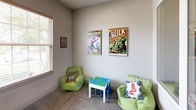 This single-floor recreation space is designed to engage and entertain, combining a playful superhero aesthetic with practical amenities. The interior is decorated throughout with bold Marvel comic book artwork, including framed prints of Spider-Man, The Hulk, and Iron Man, as well as a vivid "Wham! Boom! Zap!" pop-art canvas, lending the space a lively and dynamic character.

The children's lounge area features two green bean bag chairs accented with comic-themed throw pillows, a small activity table, and natural light filtering in through blinds-covered windows — creating a welcoming and comfortable environment for younger visitors. Adjacent to this is a dedicated gaming and media station, complete with a flat-screen TV, gaming console, and a dark wood cabinet with granite countertop, offering a well-appointed setup for interactive entertainment.

A pass-through interior window connects the lounge area to a fitness room, which is visible and accessible through a frosted French door. The fitness room is outfitted with a variety of cardio and strength equipment, well-lit by overhead lighting and additional natural light from its own windows. The neutral gray carpet and light-toned walls throughout the space provide a clean, cohesive backdrop that allows the colorful décor to take center stage.