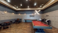 Located at Hardy Yards in Houston, TX, this single-floor amenity space is designed to serve as a refined social and recreational hub. The entry lobby sets an immediate tone with its decorative tile flooring—featuring a striking inlaid pattern combining light stone and wood-toned accents—exposed brick detailing, and large windows that invite natural light. The open lounge area beyond is furnished with eclectic seating arrangements, colorful artwork, and a pendant chandelier, creating an inviting and polished gathering space.

The game room is a standout feature of this amenity space, thoughtfully designed with layered visual interest. Deep slate-blue walls rise above wainscoting finished in crisp paneled millwork, complemented by warm wood-look tile flooring throughout. A dramatic X-shaped chandelier with exposed Edison bulbs serves as the room's focal point, illuminating a central red-felt billiards table with a rustic wood frame. A shuffleboard table lines one wall alongside a scoreboard, while a curved, tufted grey banquette with round black tables and gold-accented chairs provides comfortable lounge seating. High-top tables and barrel chairs offer additional casual seating. Wall sconces add warmth throughout, contributing to the room's upscale yet relaxed ambiance.