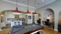This single-floor space is thoughtfully designed to blend entertainment and functionality in a seamless open-concept layout. The centerpiece of the main living area is a full-size pool table, dramatically lit by three bold red industrial-style pendant lights that hang from the crown-molded ceiling, lending a striking visual character to the room. The neutral, warm-toned walls and tile flooring create a cohesive and inviting ambiance throughout.

Flowing directly from the game area, the kitchen is well-appointed with white cabinetry, a large central island with bar seating, a decorative tile backsplash, and stainless steel appliances. The open sightlines between the kitchen and recreation area make this space ideal for entertaining.

An arched doorway leads to a dedicated workspace nook, featuring a built-in desk surface framed by an accent wall of stacked stone, adding a rustic yet refined touch. Recessed lighting and thoughtful architectural details, including arched passages and crown molding, are consistent throughout, elevating the overall design. The combination of entertainment, culinary, and work areas makes this a versatile and well-curated space suited for both leisure and productivity.