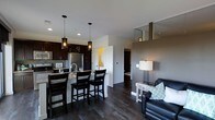 This single-floor space showcases a thoughtfully designed open-concept layout that effortlessly blends the kitchen, dining, and living areas into one cohesive environment. The kitchen is appointed with rich dark cabinetry, stainless steel appliances, and a granite-topped island that doubles as a breakfast bar, complete with dark wood bar stools. Two elegant pendant lights hang above the island, adding a warm, focused glow to the workspace. The backsplash and countertops provide tasteful contrast against the deep cabinetry tones. Adjacent to the kitchen, the dining and living areas flow naturally, anchored by wide-plank dark hardwood flooring that runs throughout the space, lending a sense of warmth and continuity. The living area features a sleek leather sofa accented with decorative pillows, accompanied by a side table and lamp that create an inviting atmosphere. Neutral gray walls provide a modern, understated backdrop that complements the darker furnishings and finishes. Recessed lighting throughout ensures consistent illumination, while a hallway visible in the background hints at additional private spaces beyond. The overall ambiance is contemporary and well-appointed, combining functional design with modern aesthetic sensibility.