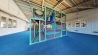 Located at 500 Hambrick Rd in Stone Mountain, Georgia, this single-floor indoor recreational facility is purpose-built for active play and community use. The centerpiece of the space is a large, multi-level soft play structure constructed with teal tubular framing, mesh safety netting, climbing elements, slides, and a distinctive circular porthole feature — offering children an engaging and dynamic play environment. The flooring throughout is covered in blue rubberized carpeting, providing a safe and cushioned surface, with additional rubber matting laid beneath the play structure for extra impact absorption. The facility features a vaulted ceiling supported by exposed steel beams, lending the space an open and airy feel with generous overhead clearance. Large floor-to-ceiling glass panels run along one side of the building, allowing abundant natural light to flood the interior and offering views of the surrounding greenery. A low rustic stone wall lines the windowed perimeter, adding a distinctive architectural character. White-paneled walls with multi-pane interior windows separate adjacent rooms, and seating benches are positioned throughout the open area for supervising adults. Signage indicating playground rules is posted near the entrance. The overall ambiance is bright, welcoming, and well-suited for recreational programming.