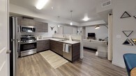 This single-floor residence is designed with a clean, contemporary aesthetic throughout. The open-concept kitchen anchors the main living area, outfitted with dark wood-grain cabinetry, stainless steel appliances, white quartz countertops, and pendant lighting above the peninsula. Wood-look flooring extends seamlessly from the kitchen into the adjacent living room, which features a tray ceiling detail and windows dressed with linen curtains, allowing natural light to filter in.

A dedicated dining area is positioned near a bank of windows, creating a bright, well-defined space finished with the same wood-look flooring. The layout includes multiple bedrooms, each carpeted for comfort. The primary bedroom is generously proportioned with windows providing natural light, while the additional bedrooms offer flexible configurations suitable for varied uses.

Two bathrooms serve the unit. The full bathroom features wood-look tile flooring, a dark vanity with white countertop, a large mirror, and a tub-shower combination. The secondary bathroom is a compact half-bath with a streamlined vanity and light flooring. Neutral gray walls are consistent throughout, contributing to a cohesive, modern ambiance across the entire space.
