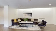 Located at 41 Cowie Hill Rd in Halifax, Nova Scotia, this building lobby offers a well-designed, single-floor space that combines functionality with modern aesthetic appeal. The entrance greets visitors with large glass doors and floor-to-ceiling glazing that floods the space with natural light, offering clear views of the surrounding tree-lined streetscape. Polished stone-effect flooring runs throughout, complemented by clean white walls and recessed lighting that creates a bright, inviting atmosphere. A prominent cylindrical structural column anchors the central hallway, adding an architectural focal point while defining the transition between the entrance and interior areas. Elevators are conveniently positioned within the hallway, ensuring easy access throughout the building. The adjacent lounge area is thoughtfully furnished with a large dark sectional sofa, an accent chair with gold-toned framing, and a white marble-top coffee table set atop a layered area rug. Subtle LED cove lighting illuminates the rear wall, highlighting a framed landscape artwork that adds warmth and character to the space. Together, these elements create a cohesive, contemporary lobby that makes a strong first impression.