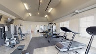 Located at 20 Pine Hall Dr in Clayton, NC, this single-story fitness center is designed to provide a comprehensive workout experience in a bright and spacious environment. The main gym area showcases vaulted ceilings that contribute to an open, airy atmosphere, with a combination of recessed and fluorescent lighting ensuring the space is well-lit throughout. The polished concrete floors are accented by interlocking rubber mat sections strategically placed under equipment for safety and stability. Large windows with blinds and a full-width glass garage-style door allow abundant natural light to fill the room while offering views of the surrounding exterior. The facility is outfitted with a diverse array of exercise equipment, including treadmills, rowing machines, cable and multi-station strength machines, adjustable benches, and additional cardio equipment. Multiple wall-mounted flat-screen televisions are positioned throughout the gym for entertainment during workouts. A wall of mirrors enhances the sense of depth and supports proper form during exercise. Adjacent to the main workout floor is a dedicated children's play area, a compact and cheerfully designed room featuring colorful foam floor tiles, a child-sized table and chairs, an abacus toy, and a wall-mounted TV — making the facility family-friendly and accommodating for parents.