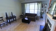 This single-floor space centers around a comfortable living room that combines functional design with an inviting ambiance. The room is anchored by a large window dressed with sheer green curtains and horizontal blinds, allowing natural light to filter in while maintaining privacy. The window offers a view of lush greenery outside, enhancing the connection to the outdoors.

The living room features neutral-toned carpeting that complements the light-colored walls, which are accented by a contrasting gray-blue feature wall behind the seating area. Crown molding adds a refined architectural detail to the space. A dark upholstered sofa serves as the focal point, paired with a tufted black ottoman that doubles as a coffee table, topped with a decorative crystal decanter set.

Additional furnishings include a dark console table along one wall and an elegant side table with a vase of dried florals, contributing to the room's sophisticated yet comfortable character. A potted plant in the corner introduces a natural element. Bold black-and-white wall art provides visual contrast, rounding out a space that feels both polished and livable.