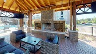 Located at 205 River Pl in Augusta, Georgia, this covered outdoor pavilion is a thoughtfully designed community gathering space that combines rustic craftsmanship with comfortable, resort-style amenities. The structure is anchored by a striking full-width brick fireplace, complete with built-in firewood storage on either side, serving as a dramatic focal point for the lounge area. The soaring pitched roof is constructed from exposed timber framing and warm wood decking, supported by substantial brick and wood columns that lend the space a strong architectural character. Ceiling fans are integrated throughout to ensure year-round comfort. The flooring throughout the pavilion is laid in a classic herringbone brick pattern, adding texture and visual interest underfoot. The open-air design allows for generous natural light and unobstructed sightlines to the surrounding pool deck and landscaped grounds beyond. Seating is arranged throughout to accommodate multiple uses, including wicker lounge sofas with navy cushions, conversation groupings, and dining sets with chairs. Mounted flat-screen televisions are positioned for visibility across seating areas. The pavilion's open sides frame views of the adjacent pool and the broader residential community, creating a seamless indoor-outdoor experience ideal for relaxation and social gatherings.