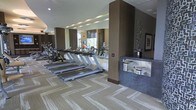 This contemporary fitness center offers a thoughtfully designed single-floor workout environment that balances functionality with upscale aesthetics. The space features a row of treadmills and cycling stations arranged for optimal use of the floor area, complemented by additional strength and cardio equipment visible throughout. Large windows along one wall invite natural light and provide views of the surrounding greenery, creating an open and energizing atmosphere. Full-length mirrors are strategically positioned to enhance the sense of space and allow exercisers to monitor their form. Structural columns clad in warm wood tones add a refined architectural element, while the patterned, neutral-toned carpet with a herringbone-inspired design lends a polished finish underfoot. A built-in water fountain station, finished in a textured blue mosaic tile, is conveniently integrated into the layout for easy access. Decorative wall art and sculptural accents contribute to the upscale ambiance of the facility. The recessed ceiling with embedded lighting ensures the space is well-lit throughout, making it a comfortable and motivating environment for a wide range of fitness activities.