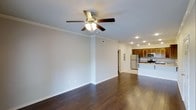 This single-floor residence offers a bright, open-concept layout that seamlessly connects the living area to the kitchen. The living space is finished with rich, dark hardwood flooring that extends throughout, complemented by crisp white walls and classic crown molding that lend an air of refined elegance. A five-blade ceiling fan with an integrated light fixture anchors the living area, providing both comfort and ambient lighting, while recessed lighting adds a modern touch throughout.

The kitchen is thoughtfully designed with warm wood cabinetry, a light-toned countertop peninsula, and stainless steel appliances including a refrigerator and microwave. The peninsula serves as a natural divider between the kitchen and living areas, ideal for casual dining or entertaining. The kitchen floor transitions to a lighter tile, creating a subtle yet clear distinction between the two spaces.

Natural light flows into the space, enhancing the airy and welcoming atmosphere. Neutral tones throughout the walls and ceilings create a versatile backdrop suitable for a variety of interior styles. A hallway visible from the main living area suggests additional rooms beyond, completing this well-appointed and functional single-level home.