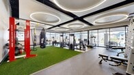 This contemporary fitness center is designed to accommodate a wide range of workout styles across three distinct exercise rooms connected by a hallway. The main exercise area is an architectural standout, featuring soaring ceilings adorned with bold black structural beams and a series of large circular LED ring fixtures that cast bright, even illumination throughout the space. Floor-to-ceiling glass walls line one side of the room, inviting abundant natural light and providing open views of the surrounding exterior. The floor transitions between rubber sport flooring and a central stretch of artificial turf, which serves as the functional training zone. This area is equipped with a heavy punching bag, TRX suspension trainers, battle ropes, and a red Octagon multi-functional rig stocked with kettlebells and accessories. Cardio and strength machines, including treadmills and cable systems, are arranged along the perimeter. A second exercise room is dedicated to cycling, featuring smart stationary bikes positioned before a full-wall mirror on light wood-style flooring, with recessed ceiling lighting creating a clean, focused atmosphere. A third room provides additional space for functional training and strength equipment, also benefiting from natural light through multiple windows. Together, the rooms form a comprehensive and well-appointed fitness facility.
