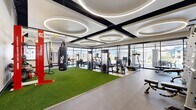 This contemporary fitness center is designed to accommodate a wide range of workout styles across three distinct exercise rooms connected by a hallway. The main exercise area is an architectural standout, featuring soaring ceilings adorned with bold black structural beams and a series of large circular LED ring fixtures that cast bright, even illumination throughout the space. Floor-to-ceiling glass walls line one side of the room, inviting abundant natural light and providing open views of the surrounding exterior. The floor transitions between rubber sport flooring and a central stretch of artificial turf, which serves as the functional training zone. This area is equipped with a heavy punching bag, TRX suspension trainers, battle ropes, and a red Octagon multi-functional rig stocked with kettlebells and accessories. Cardio and strength machines, including treadmills and cable systems, are arranged along the perimeter. A second exercise room is dedicated to cycling, featuring smart stationary bikes positioned before a full-wall mirror on light wood-style flooring, with recessed ceiling lighting creating a clean, focused atmosphere. A third room provides additional space for functional training and strength equipment, also benefiting from natural light through multiple windows. Together, the rooms form a comprehensive and well-appointed fitness facility.