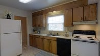 Located at 714 Riverside Drive in Salisbury, Maryland, this two-story home provides a practical and comfortable layout spread across two levels. The kitchen is a functional space featuring warm honey-toned wood cabinetry that extends to the upper and lower walls, offering generous storage throughout. A double-basin sink is positioned beneath a window fitted with white blinds, allowing natural light to filter into the space while providing a pleasant outlook. The countertops are light-colored and provide ample workspace, complementing the warm cabinetry tones. Appliances include a white electric range and a black dishwasher, creating a classic contrast within the kitchen. The flooring features light-toned vinyl, adding to the bright and clean aesthetic of the space. Crown molding along the ceiling adds a subtle architectural detail. The home's two-story configuration suggests a defined separation between living and sleeping areas, typical of traditional residential construction in the Salisbury area. Overall, the home presents a well-maintained, straightforward interior with practical finishes suited for everyday living.