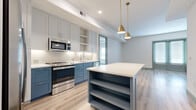 This thoughtfully designed single-floor residence combines contemporary style with functional living. The kitchen serves as the heart of the home, showcasing a striking two-tone design with crisp white upper cabinetry and bold blue lower cabinets, all complemented by clean white countertops. A generously sized central island with open shelving and a white quartz surface provides ample workspace and storage, while a pair of elegant gold pendant lights add a warm, modern touch. Stainless steel appliances complete the well-appointed cooking space. Light oak luxury vinyl plank flooring flows seamlessly throughout the entire residence, tying each room together with a cohesive, contemporary aesthetic. Sage green trim, door frames, and five-panel interior doors introduce a subtle, nature-inspired accent that enhances the overall design palette. The living area benefits from natural light admitted through full-length windows fitted with horizontal blinds, while a ceiling fan adds both comfort and style. The bedrooms are bright and well-proportioned, each featuring generous windows. The bathroom stands out with its distinctive terracotta hexagonal tile flooring, a dual-sink vanity with blue-gray cabinetry, a large frameless glass walk-in shower clad in classic white subway tile, and modern chrome fixtures throughout.