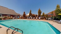 Located at 2640 Lakebridge Ln in Hilliard, Ohio, this single-level community amenity space centers around a generously sized outdoor swimming pool that serves as the focal point of the area. The pool features a classic curved design with clear, turquoise water and is surrounded by a well-maintained concrete sun deck lined with white lounge chairs, providing ample space for relaxation and sunbathing. A wrought iron-style perimeter fence encloses the pool area, ensuring both safety and a defined sense of space. Mature trees and landscaping border the fence line, offering natural shade and visual privacy from the surrounding residential buildings, which are visible in the background with their traditional pitched rooflines. The open-air setting benefits from abundant natural light under clear blue skies, creating a bright and inviting atmosphere. The overall ambiance is that of a well-appointed community retreat, designed for leisure and outdoor enjoyment within the Hilliard neighborhood setting.