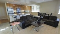 This single-floor space showcases an open-concept design that connects the kitchen, dining, and living areas into one cohesive, functional environment. The kitchen is well-appointed with warm wood cabinetry, stainless steel appliances including a microwave and range, and a granite-style countertop that extends into a breakfast bar with barstool seating — ideal for casual dining. Pendant lighting above the bar adds a welcoming touch, complementing the recessed lighting throughout. The living area features plush, dark upholstered sectional seating arranged around a sleek, modern coffee table with chrome accents, creating a comfortable and stylish gathering space. Neutral-toned carpeting grounds the living area, while the overall color palette of warm browns and beiges lends the space a cohesive, inviting ambiance. A dining area with dark wood chairs is visible adjacent to the living space, offering additional seating. A window allows natural light to filter into the kitchen area, brightening the open layout. The space is well-suited for both everyday living and entertaining, with its practical flow and modern furnishings.