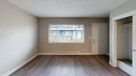This single-floor unit offers a clean, functional layout with consistent light-toned wood-style flooring and neutral warm-gray walls throughout, creating a cohesive and understated aesthetic. The living area is welcoming and filled with natural light from a large front-facing window, which also provides views of the surrounding neighborhood. A baseboard heater runs beneath the window, and a paneled entry door completes the space. The kitchen is efficiently designed with white upper and lower cabinetry, a stainless steel electric range with an overhead range hood, a stainless steel refrigerator, and a built-in sink. A striking blue subway tile backsplash adds a modern accent, while a built-in counter ledge along one wall offers additional workspace or dining space. The bedroom is a generously proportioned room with ample wall space and direct access to a private bathroom. The bathroom features a full-size soaking tub and shower combination with a curved curtain rod, complemented by clean white fixtures and brushed nickel hardware. Textured ceilings and crisp white baseboards are consistent throughout the unit, reinforcing a tidy, move-in-ready presentation. The overall layout is practical and well-appointed, suited for comfortable everyday living.