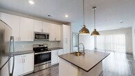 This thoughtfully designed one-bedroom, one-bathroom residence offers a cohesive open-concept layout that maximizes every square foot. The kitchen is a standout feature, outfitted with white shaker cabinetry, dark quartz countertops, a hexagonal tile backsplash, and a full suite of stainless steel appliances. A central island with seating provides additional prep and dining space, anchored by stylish pendant lighting above. The kitchen flows seamlessly into the combined living area, where large sliding glass doors invite natural light and offer views of the surrounding greenery through a private balcony. The living room is defined by a boldly colored accent wall and wood-look flooring, creating a warm yet contemporary atmosphere. The bedroom is a comfortable retreat featuring a ceiling fan, a large window with outdoor views, and plush carpet underfoot. An adjacent walk-in closet offers wire shelving along multiple walls for generous storage. The bathroom features dark wood-look flooring, white cabinetry with dark quartz countertops, a dual vanity with a wide mirror, and a glass-enclosed shower with full tile surround. The overall design aesthetic is modern and stylish, with consistent finishes tying every room together seamlessly.