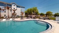 This upscale apartment complex in Melbourne, Florida, boasts a stunning resort-style pool area as its centerpiece. The expansive, curved pool features crystal-clear turquoise water and is surrounded by a spacious deck paved with light-colored stone. Multiple lounge areas are strategically placed around the pool, offering ample seating options including comfortable chaise lounges and shaded cabanas.

The pool's unique design includes a shallow area with in-water sunbeds, perfect for relaxing while partially submerged. Palm trees and other tropical plants dot the landscape, creating a lush, vacation-like atmosphere. The complex's residential buildings, visible in the background, showcase a modern architectural style with a mix of white and blue exterior finishes.

Additional amenities include a separate hot tub area, outdoor dining spaces with tables and chairs, and what appears to be a clubhouse or community building adjacent to the pool. The entire area is enclosed by a decorative metal fence, ensuring privacy and security for residents. The property's thoughtful design and attention to detail create an inviting, resort-like environment for its occupants, combining the comforts of home with the luxuries of a high-end vacation destination.