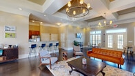 Located at 500 Carlen Ave in Lexington, SC, this single-floor community clubhouse offers a thoughtfully designed open-plan layout encompassing a living room, dining room, and kitchen. The space is defined by soaring tray ceilings adorned with detailed crown molding and anchored by a striking chandelier, lending an air of sophistication throughout. Rich, dark hardwood flooring runs seamlessly across the entire space, providing a warm and cohesive foundation.

The living area features a comfortable seating arrangement with a mix of modern and mid-century furnishings, including a tufted leather sofa, accent chairs, and a substantial wood coffee table centered on a patterned area rug. A wall-mounted flat-screen TV and a stylish media console complete the space. French doors with transom windows line the perimeter, flooding the interior with natural light and offering views of the surrounding outdoors.

The kitchen is equipped with rich wood cabinetry, granite countertops, stainless steel appliances, and a generous bar-height island with sleek blue stools—ideal for casual gatherings. The adjacent dining area accommodates a large wooden dining table with upholstered seating, bathed in natural light from plantation-shuttered windows. The overall ambiance is polished, welcoming, and well-suited for community use.