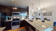 This single-floor residence offers a thoughtfully designed layout that balances functionality with contemporary style. The kitchen serves as a central highlight, showcasing rich dark cabinetry, granite countertops, a mosaic tile backsplash, and a full suite of stainless steel appliances including a refrigerator, electric range, and built-in microwave. A large island with an undermount double sink provides additional workspace and flows naturally toward the dining area, which is illuminated by an elegant overhead pendant fixture and accented by warm hardwood flooring.

The living room is generously proportioned, filled with natural light from large windows that offer outdoor views, and features neutral tones that create a welcoming atmosphere. Adjacent to it, the dining space accommodates a glass-top table beneath a statement pendant light.

The primary bedroom includes plush carpeting, natural light from a large window, and access to a spacious walk-in closet outfitted with multi-level shelving and hanging rods. A second bedroom also features carpeted flooring and ample window light. The bathroom presents granite-topped dark vanity cabinetry, hardwood-style flooring, and generous wall space. Throughout, the residence maintains a cohesive, modern aesthetic.