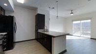 This single-floor space showcases a well-designed open-concept kitchen and living area. The kitchen is appointed with dark espresso cabinetry that extends floor to ceiling, providing generous storage. Granite countertops with subtle veining add a refined touch, while a peninsula with an integrated sink and seating overhang serves as both a functional workspace and a casual dining area. Black appliances, including a refrigerator and range, complement the dark cabinetry for a cohesive aesthetic. Two pendant lights hang above the peninsula, offering focused task lighting, while recessed lighting throughout ensures the space remains well-lit. Light-toned hardwood-style flooring runs continuously from the kitchen into the adjacent living area, creating a seamless visual flow. The living area benefits from a ceiling fan and access to natural light through a sliding glass door or large window, brightening the overall space. White walls provide a clean, neutral backdrop that contrasts effectively with the dark kitchen elements, lending the interior a contemporary, balanced character throughout.