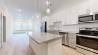 This well-appointed single-floor unit showcases a clean, contemporary design with a bright and airy aesthetic throughout. The open-concept kitchen serves as the focal point, featuring a large central island topped with granite countertops and illuminated by stylish pendant lights. Crisp white upper cabinetry is complemented by wood-toned lower cabinets, while a classic subway tile backsplash ties the design together. Stainless steel appliances, including a refrigerator, range, microwave, and dishwasher, complete the fully equipped kitchen. Light hardwood flooring flows seamlessly through the main living areas, enhancing the sense of openness and continuity. The living space is bathed in natural light from multiple windows fitted with horizontal blinds. The bedrooms offer plush carpeting underfoot and ceiling fans for comfort, with windows that bring in additional natural light. The bathroom is thoughtfully designed with granite-topped white vanity cabinetry, a soaking tub, and light hardwood flooring, along with access to a walk-in closet. Throughout the unit, neutral wall tones, recessed lighting, and modern finishes create a cohesive, move-in-ready environment that balances style and functionality.