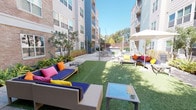 This outdoor space is part of a multi-story residential community that blends two distinct architectural styles. One section showcases a classic brick exterior with pitched rooflines, bay windows, and mature trees, creating a traditional, grounded aesthetic. The other features a more contemporary design with light-toned cladding, large windows, and clean-lined balconies.

The grounds are thoughtfully designed for outdoor living and social gathering. A resort-style swimming pool serves as a central amenity, surrounded by a paved brick deck lined with blue lounge chairs and shaded umbrella tables — ideal for relaxing or entertaining. Mature trees and manicured shrubs provide natural privacy and greenery around the pool area.

A separate courtyard offers a vibrant gathering space with artificial turf lawn, string lights overhead, and an eclectic mix of navy and rattan sectional seating accented by bold, colorful cushions in pink, orange, and yellow. Additional dining and seating clusters with umbrella shade structures are distributed throughout the paved walkways connecting the buildings.

Well-maintained landscaping, including ornamental grasses, tropical plants, and established trees, ties the outdoor areas together, creating an inviting and lively atmosphere throughout the community grounds.