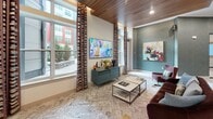 Located at 2031 Maywill St in Richmond, Virginia, this thoughtfully curated amenity space combines modern design with warm, inviting elements to create a dynamic environment suited for both relaxation and productivity. The single-floor layout is unified by a striking warm wood plank ceiling that adds richness and depth throughout, complementing the patterned carpeted flooring below.

The main lounge area is a standout, featuring floor-to-ceiling windows that bathe the room in natural light while offering views of the surrounding neighborhood. Plush velvet seating in deep burgundy tones is arranged around a sleek coffee table, with a wall-mounted flat-screen TV and a teal credenza serving as a functional focal point. Bold, colorful abstract artwork adorns the teal textured accent walls, adding a vibrant artistic character to the space.

A dedicated workspace area features a long white countertop desk illuminated by pendant lighting, set against a herringbone-tiled accent wall — ideal for focused work or casual tasks. Nearby, elegant wood slatted room dividers provide subtle spatial definition without fully enclosing areas, maintaining an open and airy flow throughout.

Glass-paneled doors lead between zones, enhancing transparency and connectivity. The overall design palette — combining teal, warm wood, cream, and rich jewel tones — creates a cohesive, sophisticated ambiance that is both functional and aesthetically refined.