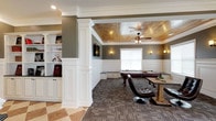 This single-floor community space is designed with a sophisticated yet welcoming aesthetic, combining classic architectural elements with comfortable, functional furnishings. Throughout the space, white wainscoting, crown molding, and decorative columns establish a consistent, polished character, while neutral taupe-gray walls provide a refined backdrop.

The game room stands out as a focal point, featuring a tray ceiling clad in warm wood planking, complemented by a ceiling fan and wall sconces. A full-sized billiards table anchors the space, which is finished with patterned carpet tiles. Built-in shelving and bench seating along the walls add both storage and charm.

Adjacent to the game room, a comfortable lounge area offers upholstered seating arranged around a small accent table, with natural light filtering through multiple windows. Rich hardwood flooring flows through the lounge and a separate dining area, where leather sofas and dining tables with chairs provide ample seating for gatherings. A wall-mounted television enhances the lounge.

The kitchen features white cabinetry, dark granite countertops, black appliances, and a curved breakfast bar with bold red barstools. Pendant lighting adds warmth overhead. A diagonal-tiled corridor connects the various areas, enhancing the sense of flow and spaciousness throughout.