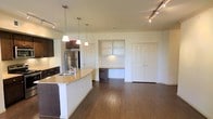 Located in Midland, this single-floor space offers a thoughtfully designed open-concept layout that seamlessly connects the kitchen and living areas. The kitchen is a standout feature, showcasing dark espresso cabinetry, stainless steel appliances including a full-size refrigerator, and a granite-topped island with a double sink. Three elegant pendant lights hang above the island, adding a warm, focused glow to the workspace. Rich hardwood flooring runs throughout, grounding the space with a sense of warmth and continuity. Track lighting along the ceiling provides additional illumination across the open living area, which benefits from a bright, airy atmosphere accentuated by light-toned walls. A built-in desk nook with open shelving is integrated into one wall, offering a practical and dedicated workspace without sacrificing floor space. A set of double doors provides generous closet storage nearby. Crown molding details along the ceiling add a refined architectural touch, elevating the overall finish of the space. The combination of functional built-ins, modern fixtures, and cohesive design elements makes this a well-appointed and versatile single-level space suitable for a variety of lifestyles.