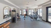 Located at 6354 W Longboat Blvd in Tampa, FL, this fitness center is a well-appointed, single-floor facility designed to support a comprehensive workout experience. The space features a clean, modern aesthetic with neutral-toned walls, textured accent panels, and durable carpet tile flooring throughout. Ceiling fans and recessed lighting provide consistent airflow and illumination across the room.

The cardio zone is equipped with multiple treadmills and stationary bikes, positioned near large windows that offer views of the surrounding exterior, allowing natural light to fill the space. A dedicated strength training area houses a selection of Precor selectorized machines, including cable systems and resistance stations with padded amber-toned benches, offering a variety of upper and lower body exercises.

A free weight section features a fully stocked dumbbell rack alongside a weight bench, catering to users who prefer traditional lifting. An arched decorative mirror enhances the visual depth of this zone while maintaining a polished appearance. The facility includes clearly marked exit doors for safety and is accessible via glass-paneled entry doors. Overall, the space strikes a balance between functionality and modern design, creating an inviting environment for fitness.
