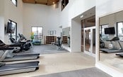 Located at 986 Broadway in Chula Vista, this fitness center combines striking architectural design with a well-appointed selection of exercise equipment. The space immediately impresses with its soaring ceilings adorned with a warm wood slat panel design and recessed lighting, creating an open, airy atmosphere that sets it apart from a typical gym environment. Crisp white walls amplify the sense of volume and allow natural light from strategically placed windows to fill the space throughout the day. The main workout floor features a row of Matrix treadmills and cardio machines for aerobic training, complemented by elliptical trainers and stationary bikes. A dedicated free weights area lines one wall, equipped with a comprehensive dumbbell rack and floor-to-ceiling mirrors that visually expand the space while allowing users to monitor their form. A wall-mounted television keeps members entertained during workouts. Toward the rear of the floor, a storage unit houses exercise balls, medicine balls, and additional fitness accessories for functional training. The flooring is a durable, cushioned surface well-suited for high-traffic fitness use. The overall ambiance is clean, modern, and motivating, making this fitness center a polished and practical facility for residents and users of the Broadway location.