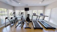 The Village Fitness Center is a thoughtfully designed, single-floor facility that provides residents with a comprehensive workout environment. The space is accessed through a set of distinctive black-framed glass sliding doors, offering a clear sightline into the gym before entry. A water cooler is conveniently positioned in the entryway area for easy access.

The main workout floor features polished concrete flooring throughout, lending the space a clean, durable aesthetic. Large windows with horizontal blinds line multiple walls, inviting abundant natural light and providing views of the surrounding greenery. A ceiling fan and recessed lighting ensure the space remains well-ventilated and adequately lit at all times.

Cardio equipment is well-represented, with multiple treadmills, elliptical trainers, and a recumbent stationary bike arranged to take full advantage of the natural light and outdoor views. A wall-mounted flat-screen television is positioned centrally for entertainment during workouts.

A dedicated strength training area includes a multi-station cable machine suitable for a variety of resistance exercises, as well as a full rack of dumbbells in graduated weights. A large floor-to-ceiling mirror spans one wall, visually expanding the space and allowing users to monitor their form. The textured ceiling adds a finishing touch to this practical and inviting fitness facility.