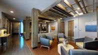 This single-floor space showcases a compelling fusion of rustic and contemporary design. The most striking architectural feature is the exposed wooden beam ceiling structure, supported by substantial wood-clad columns that add warmth and an industrial character to the interior. The beams are finished in a natural, weathered tone, creating a strong visual contrast against the smooth, light-painted walls and recessed lighting.

The lounge-style seating area is thoughtfully arranged, featuring modern upholstered armchairs in neutral grey and blue tones, complemented by accent cushions in muted yellow-green. A low, round side table topped with a decorative metallic centerpiece anchors the central seating grouping. The patterned carpet in warm terracotta and brown tones adds richness underfoot and defines the lounge zone.

Along the perimeter, solid wood doors with paneled detailing suggest access to additional rooms or facilities, including an accessible room indicated by visible signage. A piece of abstract wall art in warm ochre tones adds a curated finishing touch. Recessed and track lighting ensures the space is well-illuminated throughout, contributing to a welcoming and polished ambiance.