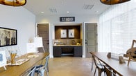 This single-floor studio is designed with creativity and functionality in mind. The space features polished concrete floors that lend an industrial-chic aesthetic, complemented by crisp white walls and recessed lighting that ensures consistent, even illumination throughout. A prominent "STUDIO" sign anchors the focal wall, which houses a built-in dark cabinetry wet bar complete with a sink and counter space — practical for art supply prep or light refreshments. The nook is framed by a richly textured dark accent wall, adding depth and visual contrast to the otherwise light and airy interior. Two classic panel doors in a muted gray tone flank the central wet bar area, providing access to adjacent rooms or storage. Large windows along the side walls invite generous natural light, enhancing the open and welcoming ambiance. The main area is furnished with wooden tables and metal bistro-style chairs, creating a versatile workspace suited for classes, workshops, or collaborative sessions. An artist's easel stationed near the entrance further underscores the studio's creative purpose. Overall, the space strikes a balance between modern design and practical utility, making it a well-appointed environment for artistic endeavors.