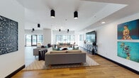 Located at 355 N Rosalind Ave in the heart of Orlando, FL, this contemporary single-floor amenity space offers an expansive, open-plan layout designed for both relaxation and socializing. Throughout the space, warm-toned hardwood-style flooring provides a cohesive foundation, complemented by crisp white walls and bold black base molding.

The main lounge area is anchored by multiple seating arrangements featuring plush gray and teal upholstered furniture, area rugs, and a large wall-mounted flat-screen television. Sleek cylindrical pendant lights and geometric linear fixtures illuminate the space, adding a modern industrial touch to the bright, airy atmosphere.

A dedicated games area includes a full-size billiards table and additional casual seating clusters, ideal for informal gatherings. Floor-to-ceiling windows and glass-panel doors line one side of the space, flooding the interior with natural light and providing direct views of an outdoor area beyond.

The kitchen zone features an elongated run of light gray cabinetry with white countertops, a mosaic tile backsplash, and a central island with bar-height seating and pendant lighting overhead. Curated artwork adorns the walls throughout, adding color and character. The overall ambiance is modern, welcoming, and well-appointed for community use.