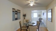This single-story space offers a comfortable and functional layout with a warm, understated aesthetic. The dining area serves as a central feature, accommodating a round glass-top table surrounded by upholstered chairs with dark wood frames — an inviting setup for everyday meals or casual gatherings. Hardwood flooring runs throughout, adding warmth and continuity to the space. A ceiling fan with a built-in light fixture is centrally mounted, providing both airflow and ambient lighting. The walls are painted in a neutral white tone, creating a clean backdrop that is complemented by decorative wall art in muted, abstract tones. A tall decorative vase with dried botanicals adds a natural accent to the corner. An open passageway connects the dining area to an adjoining room, which benefits from abundant natural light streaming through a window, creating a bright and airy feel. The secondary space appears to offer additional seating or utility, enhancing the overall flow of the layout. Baseboard heating is visible, indicating practical year-round comfort. Overall, the space presents a simple, well-maintained interior with a neutral palette suitable for a variety of styles.
