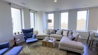 This single-floor space showcases a thoughtfully designed living area that blends contemporary style with comfortable functionality. The living room is a standout feature, benefiting from multiple large windows that flood the interior with natural light while framing elevated views of the surrounding urban cityscape, suggesting the unit is positioned on an upper floor of a larger building.

The room's design follows a neutral color palette, with light gray walls providing a clean, modern backdrop. A generously sized L-shaped sectional sofa in a warm beige tone serves as the centerpiece, complemented by a bold blue wingback chair with a matching ottoman that introduces a deliberate pop of color. Decorative throw pillows in navy and white geometric patterns tie the two seating areas together cohesively.

A slatted wood coffee table anchors the seating arrangement atop a textured area rug, adding warmth and organic contrast to the otherwise contemporary setting. Recessed ceiling lighting supplements the abundant natural light. The smooth hardwood flooring visible at the room's edges adds to the polished, urban aesthetic of the overall space.