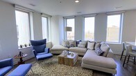 This single-floor space showcases a thoughtfully designed living area that blends contemporary style with comfortable functionality. The living room is a standout feature, benefiting from multiple large windows that flood the interior with natural light while framing elevated views of the surrounding urban cityscape, suggesting the unit is positioned on an upper floor of a larger building.

The room's design follows a neutral color palette, with light gray walls providing a clean, modern backdrop. A generously sized L-shaped sectional sofa in a warm beige tone serves as the centerpiece, complemented by a bold blue wingback chair with a matching ottoman that introduces a deliberate pop of color. Decorative throw pillows in navy and white geometric patterns tie the two seating areas together cohesively.

A slatted wood coffee table anchors the seating arrangement atop a textured area rug, adding warmth and organic contrast to the otherwise contemporary setting. Recessed ceiling lighting supplements the abundant natural light. The smooth hardwood flooring visible at the room's edges adds to the polished, urban aesthetic of the overall space.