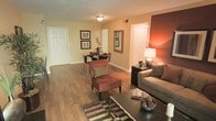 This single-floor residence showcases an open-concept layout that seamlessly connects the living and dining areas, creating a welcoming and functional flow throughout. Rich hardwood flooring runs continuously across the space, adding warmth and visual cohesion to the interior.

The living area is anchored by a neutral-toned sofa dressed with decorative pillows, complemented by a plush burgundy accent chair and a dark wood coffee table. A bold terracotta-toned accent wall adds depth and a warm focal point to the room, enhanced by a tasteful table lamp that casts a soft, ambient glow.

The dining area sits adjacent to the living space, accommodating a compact table and chairs, framed wall art, and decorative floral accents that contribute to the room's polished aesthetic. A mirrored wall piece reflects light and adds a sense of dimension to the space.

The entry door is clearly visible from the main living area, offering a straightforward and convenient layout. Neutral beige walls throughout provide a versatile backdrop, while flush-mount ceiling lighting ensures consistent illumination across the open floor plan.