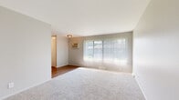 This single-floor unit offers a practical and well-organized layout designed for comfortable everyday living. The spacious living area is carpeted and bathed in natural light streaming through a large picture window fitted with vertical blinds. A wall-mounted air conditioning unit provides climate control, while a flush-mount ceiling fixture adds overhead lighting. The living space flows into an adjacent hallway with warm hardwood-style flooring, connecting the various areas of the unit seamlessly.

The bedroom is generously sized, featuring soft carpeting, a window with vertical blinds that allows for ample natural light, and a closet with a door for convenient storage. The neutral gray-toned walls throughout create a calm, cohesive aesthetic.

The kitchen is efficiently designed with white shaker-style cabinetry, white countertops, and a stainless steel sink. Black appliances — including a gas range, overhead range hood, and a full-size refrigerator — provide a striking contrast against the lighter finishes. Warm hardwood-style flooring completes the kitchen's clean and functional character.

The bathroom features a full-size soaking tub with a white tiled surround and a towel bar, maintaining the unit's consistent clean and bright aesthetic throughout.