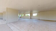 Located at 600 E Marble St in Wylie, TX, this generously sized single-floor space offers a versatile and open layout ideal for a range of uses. The interior is defined by its clean, neutral palette, with white horizontal shiplap-style wall paneling and light-toned large-format tile flooring that runs throughout, lending a bright and cohesive aesthetic.

The expansive main living area benefits from an abundance of natural light, delivered through multiple oversized windows positioned along the walls. These windows frame views of the surrounding mature tree canopy, creating a pleasant connection to the outdoors. The smooth, flat ceilings with recessed lighting contribute to the airy, uncluttered feel of the interior.

Structural columns punctuate the open floor plan, providing architectural definition without compromising the sense of spaciousness. A standout feature of the space is the expansive covered outdoor terrace, accessible from the main interior and enclosed by sleek black wrought iron railings. This open-air area is equally generous in scale, offering panoramic views of the surrounding greenery and neighborhood, making it well-suited for gatherings or events.

A brick accent column adds a contrasting textural detail to the terrace area, complementing the predominantly white interior finishes. The combination of the bright interior, seamless indoor-outdoor flow, and substantial square footage makes this a highly adaptable and visually appealing space.