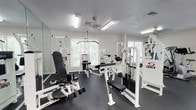 The fitness center is a bright, single-floor space designed to support a comprehensive strength training routine. The room is outfitted with a selection of Cybex brand weight machines finished in a crisp white frame with black upholstery, lending the space a clean, professional appearance. Multiple windows along the walls allow natural light to filter in, creating an open and energizing atmosphere. The dark rubber flooring provides a durable, functional surface suitable for heavy equipment and active use. A ceiling fan and overhead fluorescent lighting ensure the space remains well-ventilated and evenly lit throughout the day. Large mirrors are positioned along one wall, a practical feature for monitoring form during workouts. The textured ceiling and neutral grey-white walls keep the overall design simple and focused. The arrangement of the equipment allows for comfortable circulation between machines, making the space feel organized and accessible. Whether used for targeted muscle training or a full-body workout, this fitness center provides a well-maintained and purposeful environment for residents to stay active.