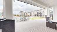 This expansive outdoor amenity area in Athens, Alabama, is designed to deliver a resort-style experience within a modern residential community. At the heart of the space is a generously sized swimming pool with a zero-entry section, surrounded by wide paver pool decks lined with rows of contemporary chaise lounges and teal umbrellas, providing ample space for sunbathing and relaxation.

A striking farmhouse-inspired clubhouse anchors the space, featuring board-and-batten white siding, a steeply pitched dark shingle roof, and arched steel-framed windows that add architectural character. An expansive covered outdoor patio extends from the clubhouse, supported by white columns and brick piers, and equipped with large ceiling fans, recessed lighting, a wall-mounted TV, and comfortable lounge seating — creating a shaded gathering space that remains inviting in any weather.

Adjacent to the pool deck, vibrant green turf lawn areas provide open space for outdoor activities. Lush landscaping beds with young plantings line the perimeter, complemented by black wrought-iron fencing that defines the amenity zone while maintaining open views of the surrounding multi-story residential buildings. The cohesive design palette of white, dark gray, and natural stone tones unifies the entire outdoor environment.