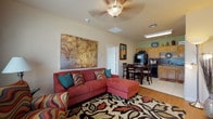 Located at 3701 Cimarron Blvd in Corpus Christi, Texas, this single-floor unit offers a well-organized layout that connects the living, dining, and kitchen areas in an open and functional arrangement. The living room is a vibrant space anchored by a bold crimson accent wall and two windows that invite natural light. Wood-look flooring, a ceiling fan, and a decorative botanical wall canvas contribute to the room's warm character. The kitchen features warm oak cabinetry, dark countertops with a tile backsplash, black appliances including a range, microwave, and refrigerator, and light tile flooring — providing ample storage and prep space. A dining area with a table and chairs sits adjacent to the kitchen, creating a convenient flow for everyday living. The bedroom offers plush carpeting, a ceiling fan, and a window with outdoor views, providing a quiet and comfortable retreat. A decorative sunburst mirror adds a stylish focal point to the room. The bathroom includes a tub-shower combination, wood-look flooring, oak cabinetry, and direct access to a walk-in closet with built-in shelving. Overall, the unit presents a colorful and well-appointed interior suited for comfortable residential living in the Corpus Christi area.